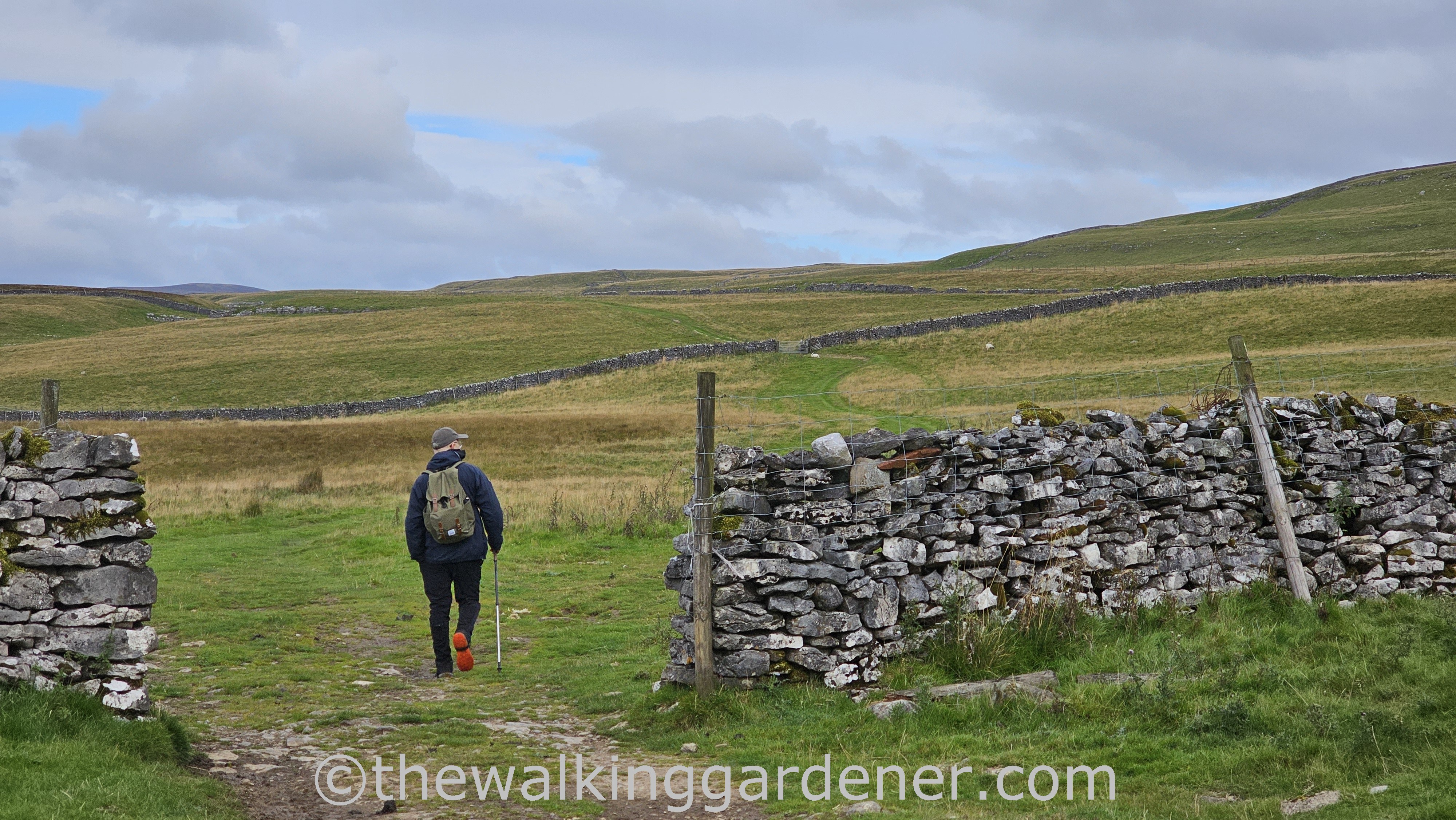 A hiker with a backpack walking through a stone gate, surrounded by picturesque green fields and dry stone walls under a cloudy sky.