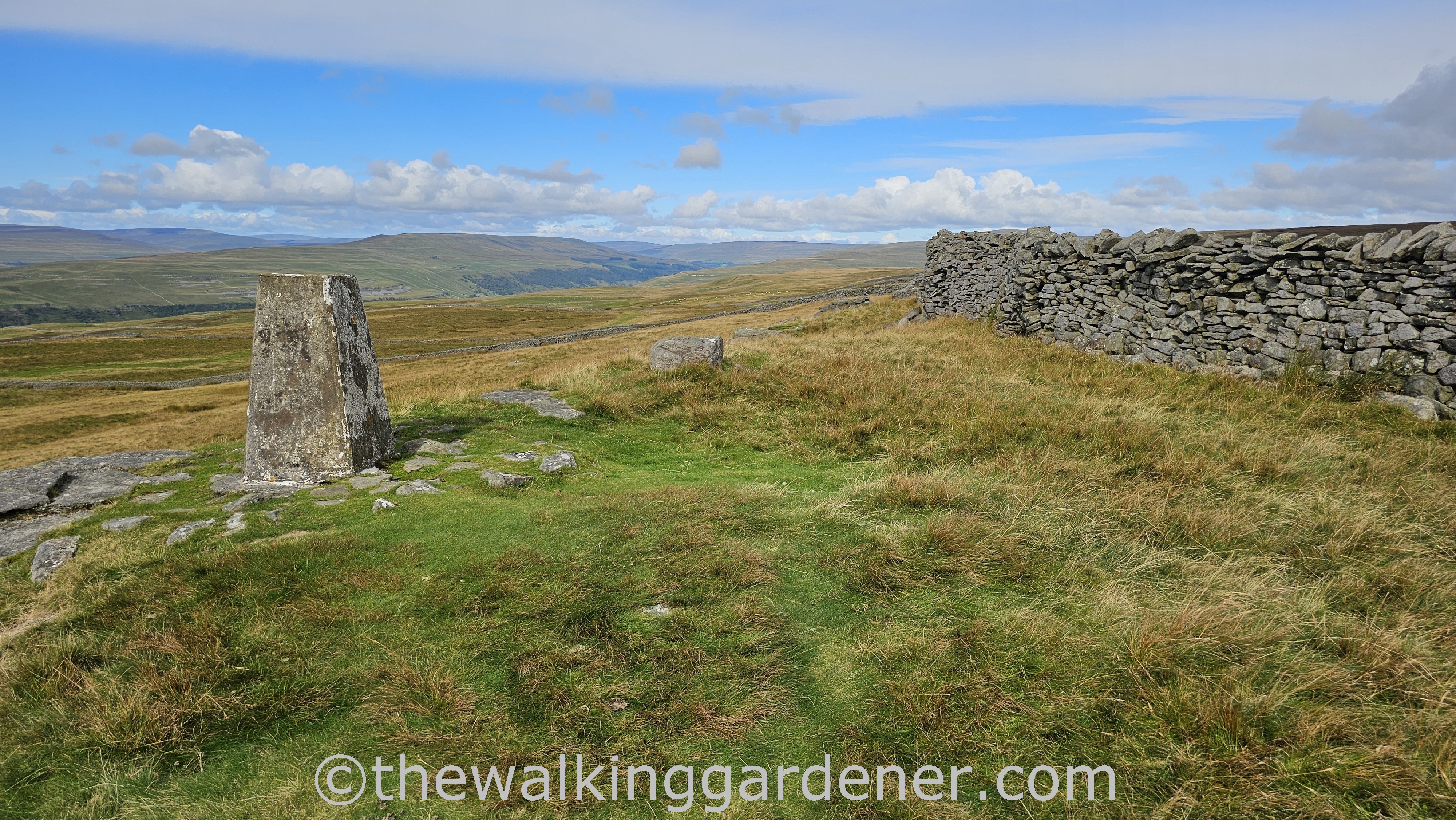 Capplestone gate trig point stands in a grassy field with drystone walls in the background, overlooking a wide landscape under a partly cloudy sky.