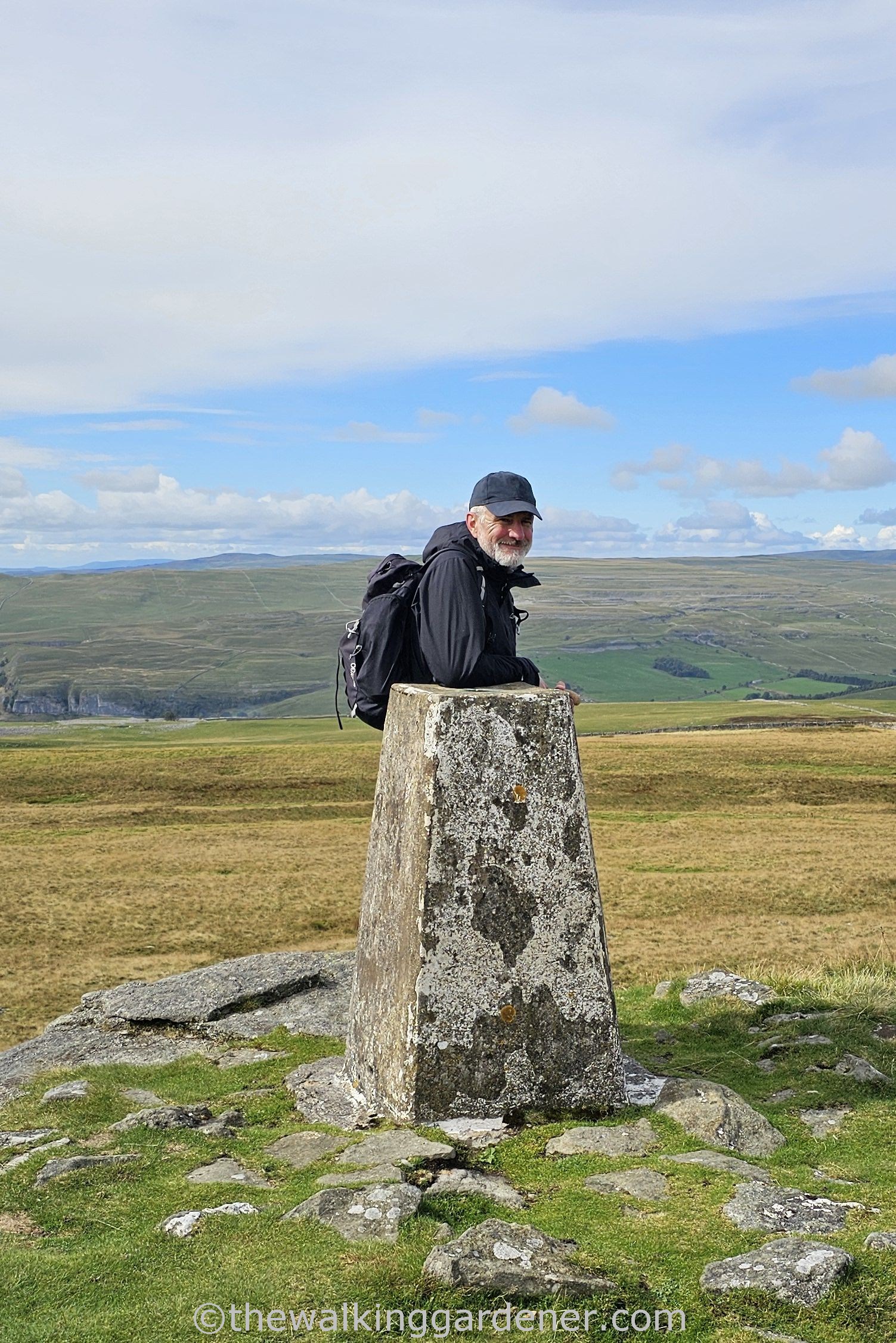 David Marsden leans against Capplestone Gate trig point on a hilltop, with green hills and a blue sky in the background.