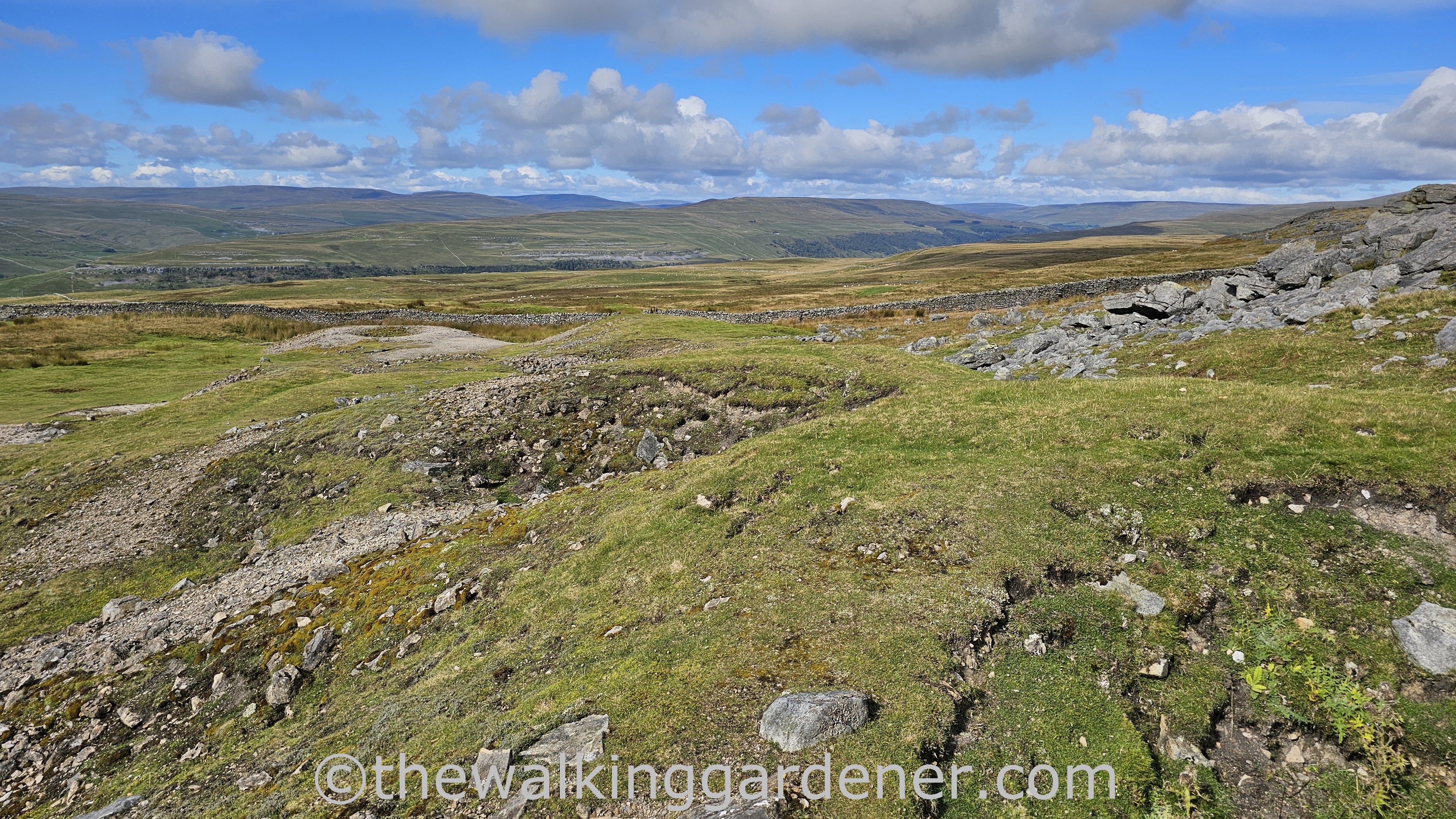 Scenic view of open grassy fell with drystone walls and distant rolling hills under a partly cloudy blue sky.