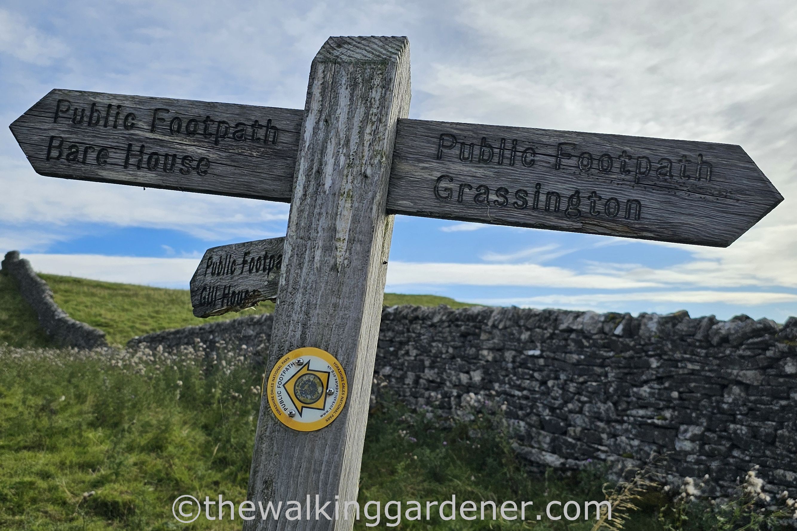 A wooden signpost indicating public footpath directions for Bare House and Grassington against a backdrop of green grass and stone walls.