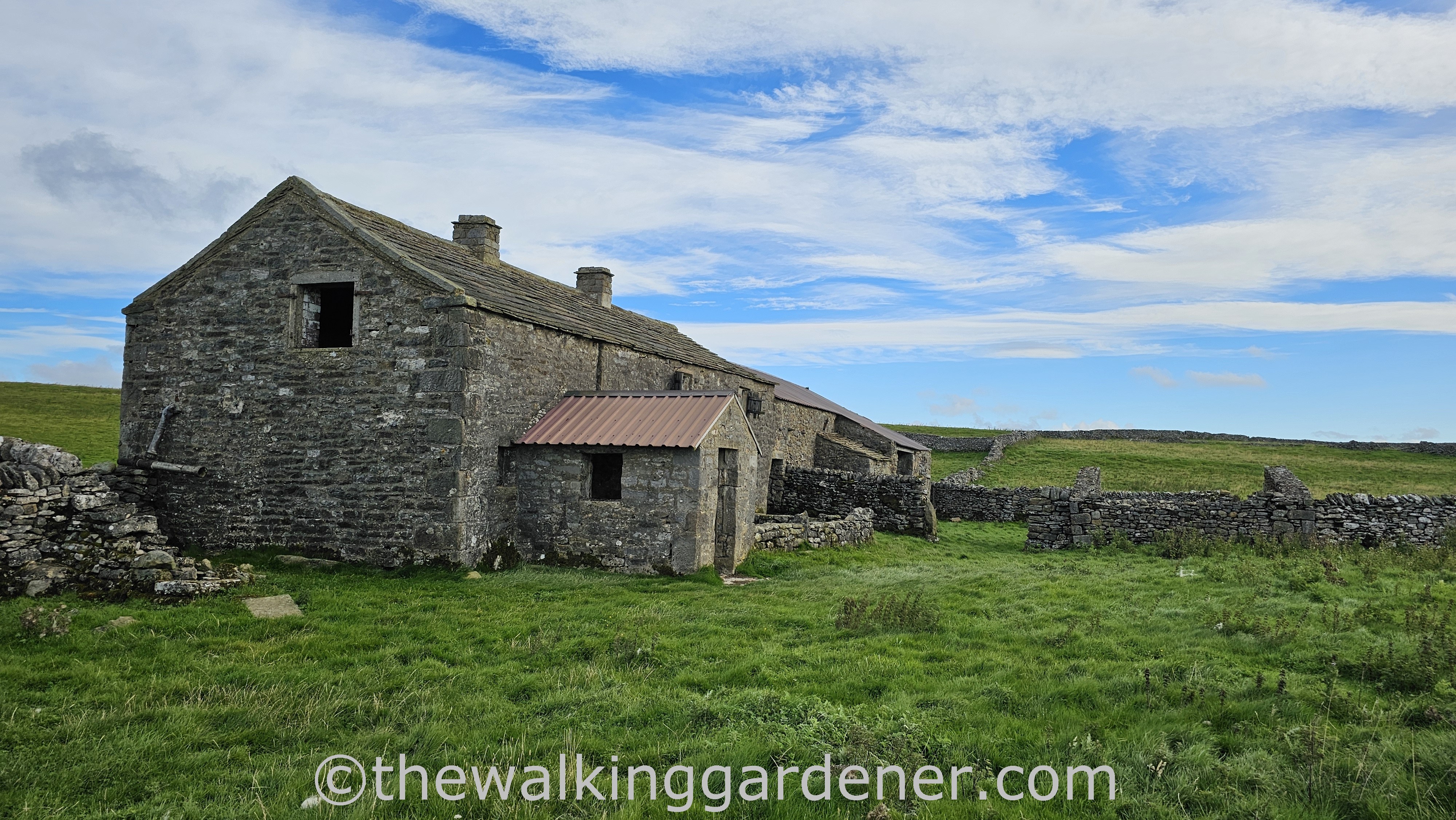 A partially dilapidated stone farmhouse - Bare House, Grassington - situated in a green pasture, with dry-stone walls visible in the background against a blue sky with fluffy clouds.