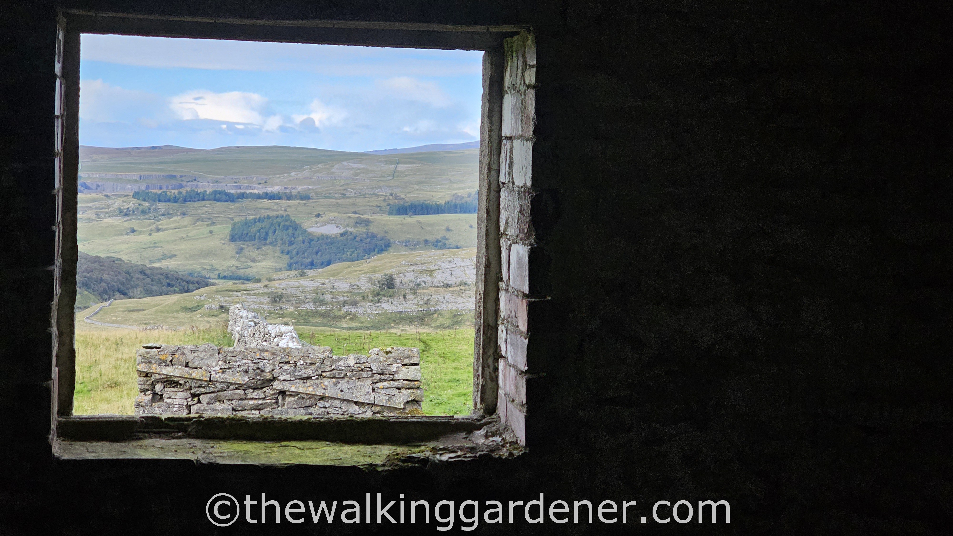 View of a green hilly landscape framed by a dilapidated window in Bare House, Grassington.