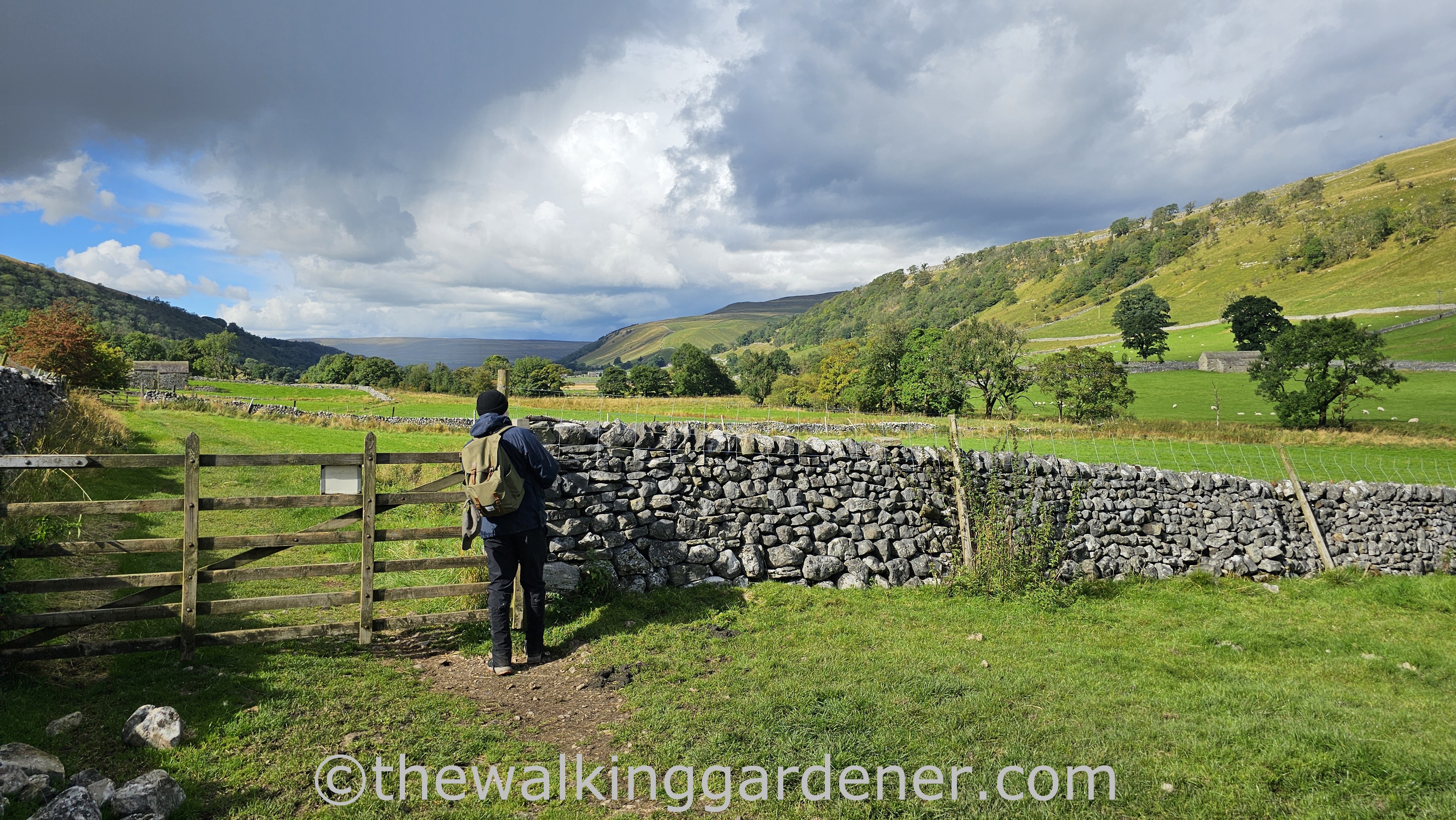 A hiker standing by a wooden gate, overlooking a picturesque landscape with lush green fields and hills under a partly cloudy sky.