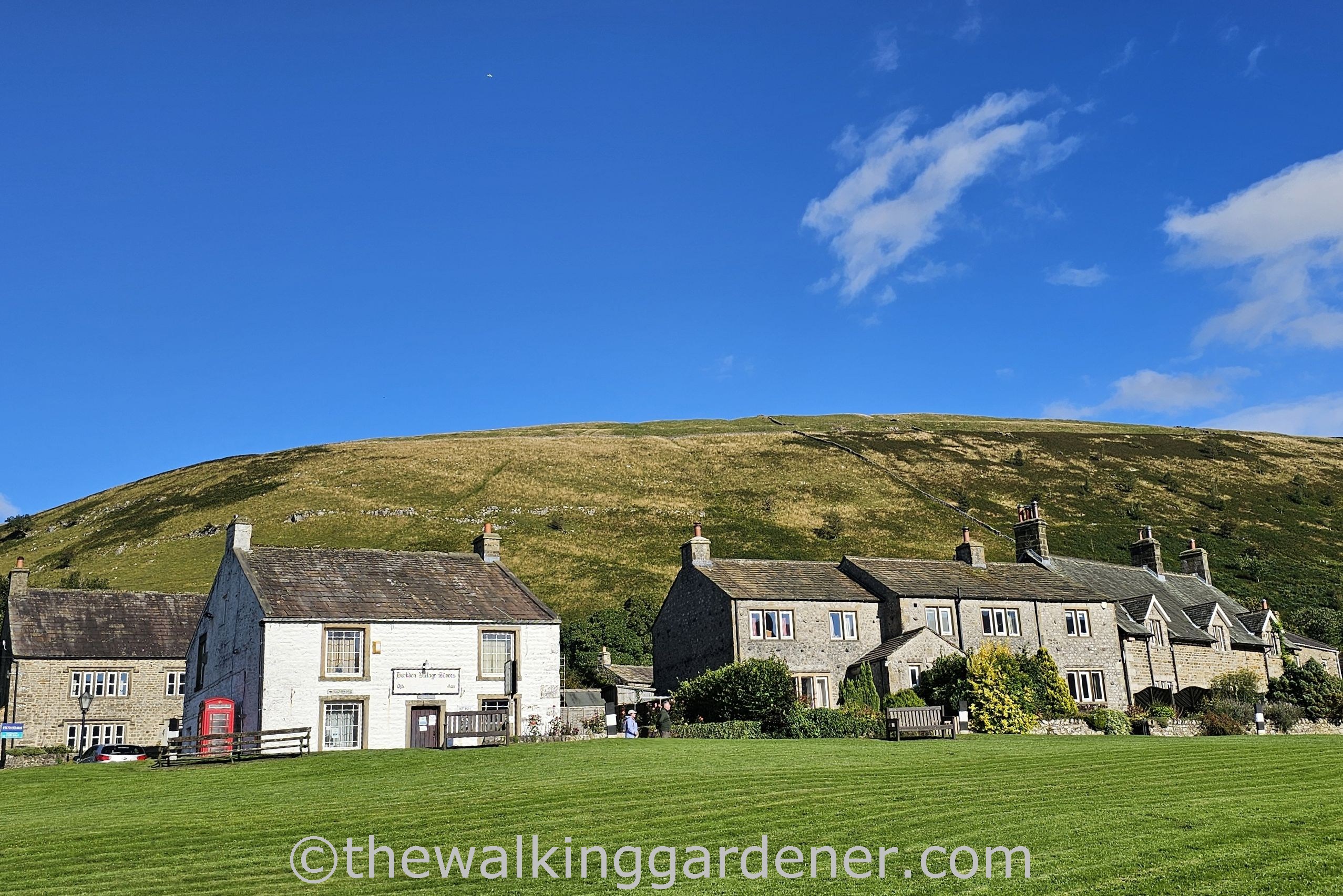 A scenic view of a small village with stone houses and a green lawn, set against a backdrop of a rolling hill and a blue sky with scattered clouds.
