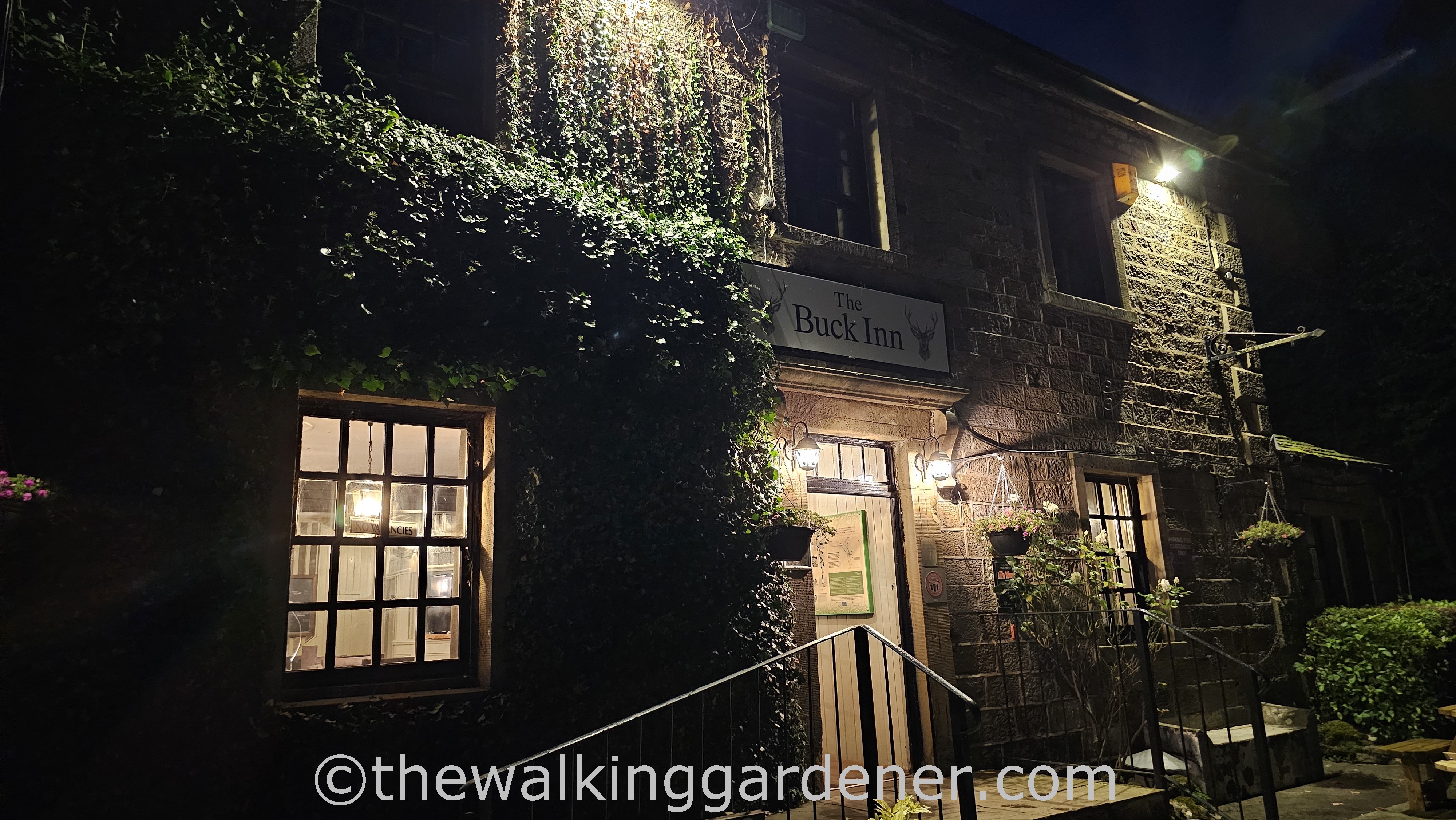 A nighttime view of The Buck Inn, Buckden - a stone building adorned with climbing plants, illuminated by warm lights.