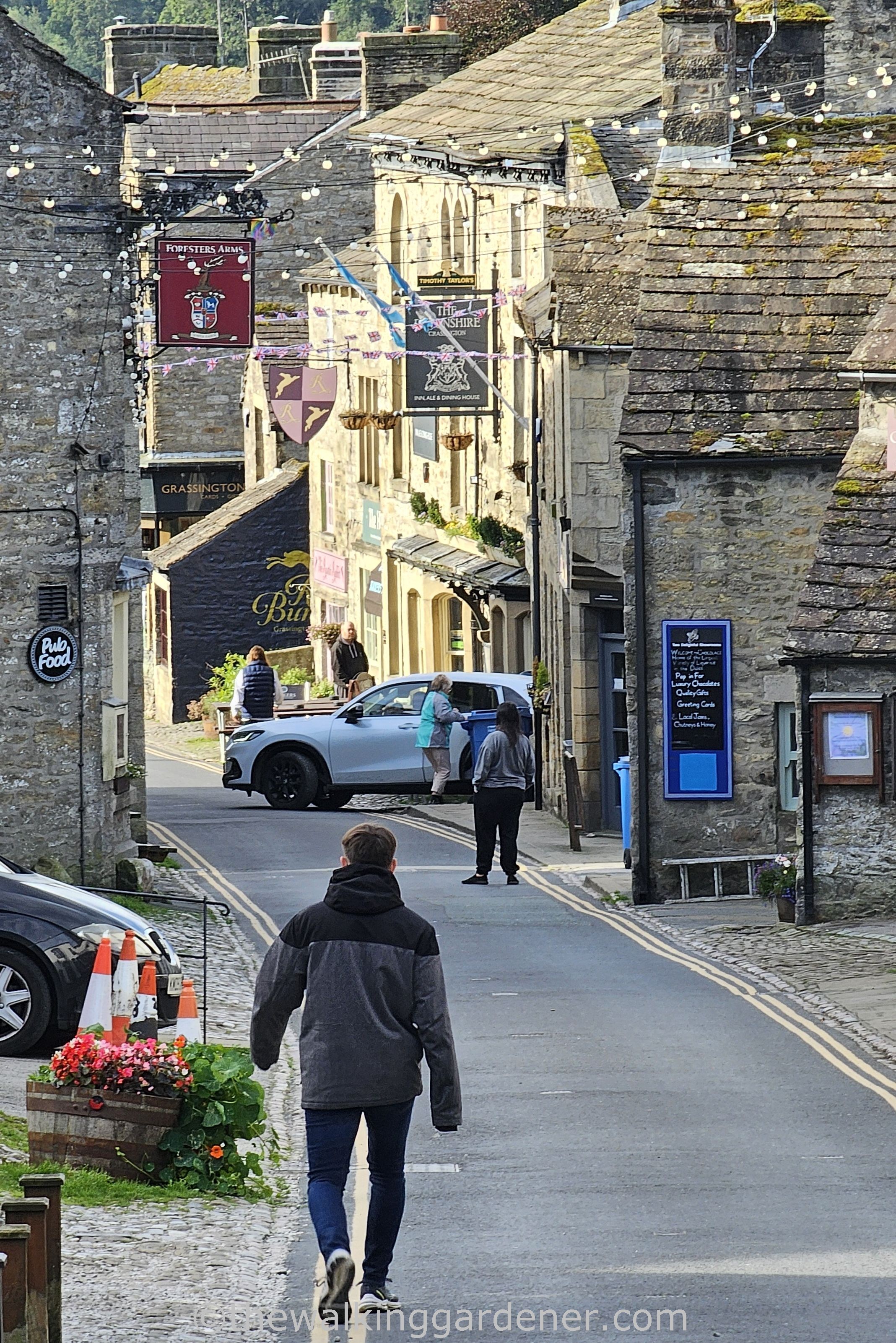 A man walking down a cobbled street in Grassington, with shops and a parked car in the background.