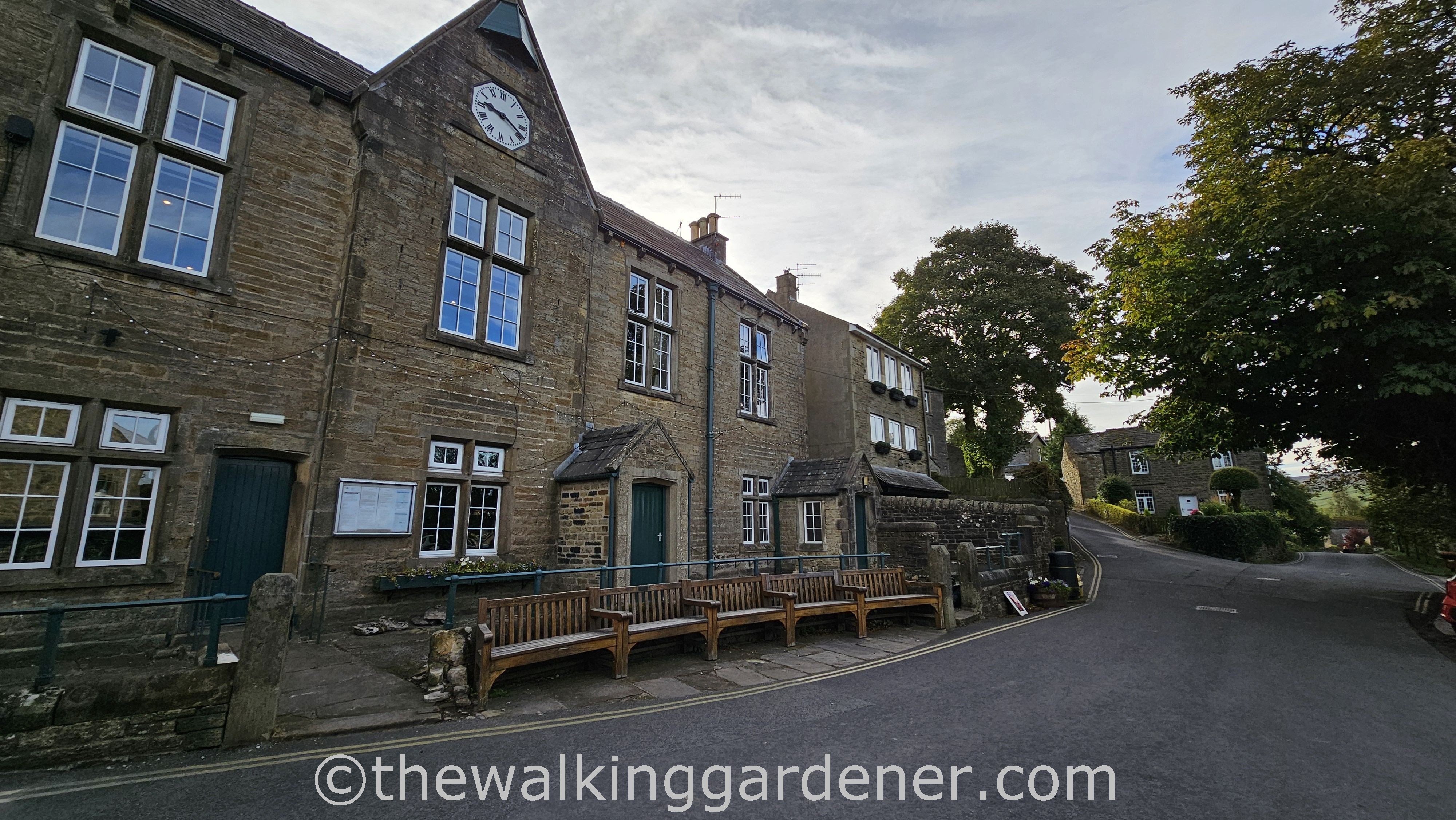 A historic stone building with large windows and a clock on the facade in Grassington, surrounded by benches and trees along a quiet street in a small town.