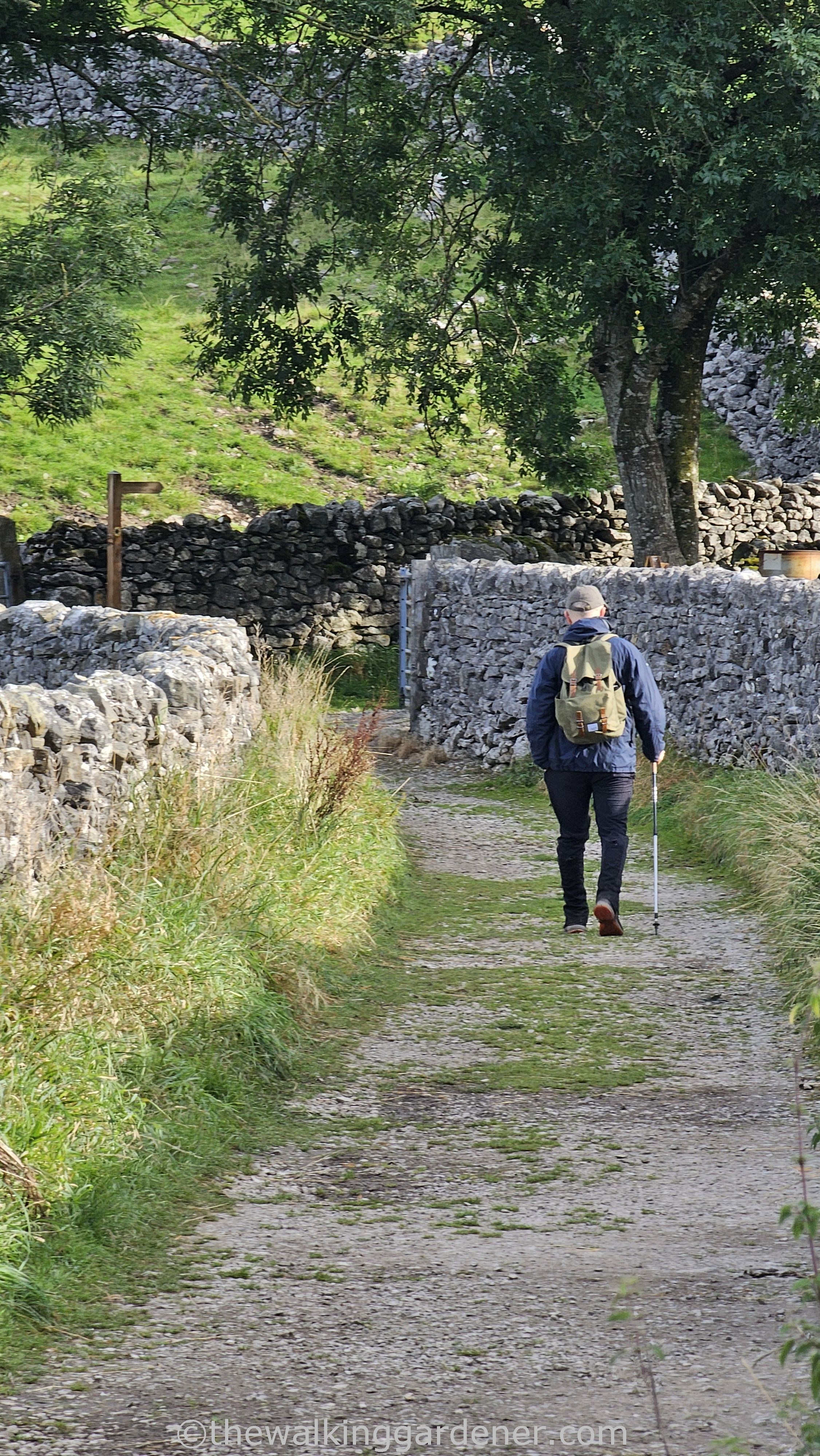 Jim Pilston walking along a gravel path in a rural area, flanked by dry stone walls and grass. The individual is using a hiking pole and carrying a backpack.