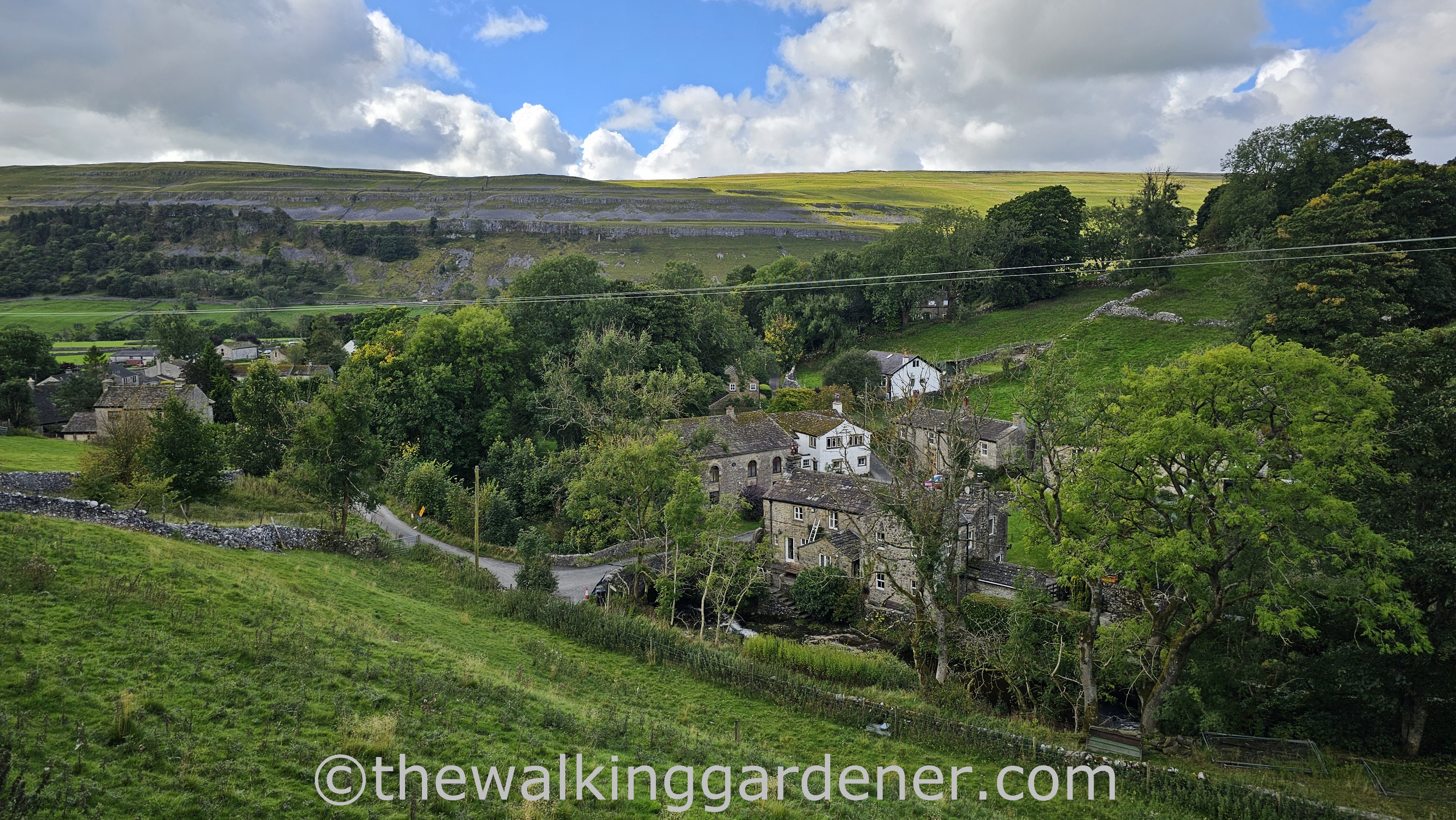 A scenic view of Kettlewell - a small village nestled in a green valley, surrounded by rolling hills and blue skies with clouds.